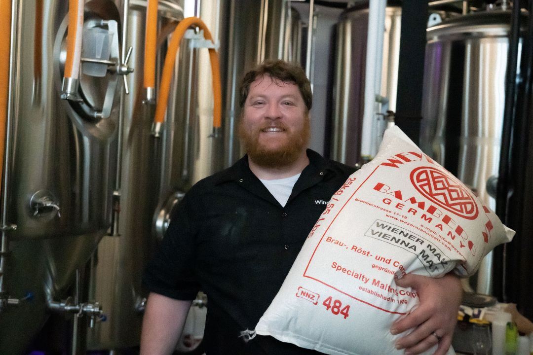 Smiling brewer holding a large malt bag inside a brewery with stainless steel fermentation tanks