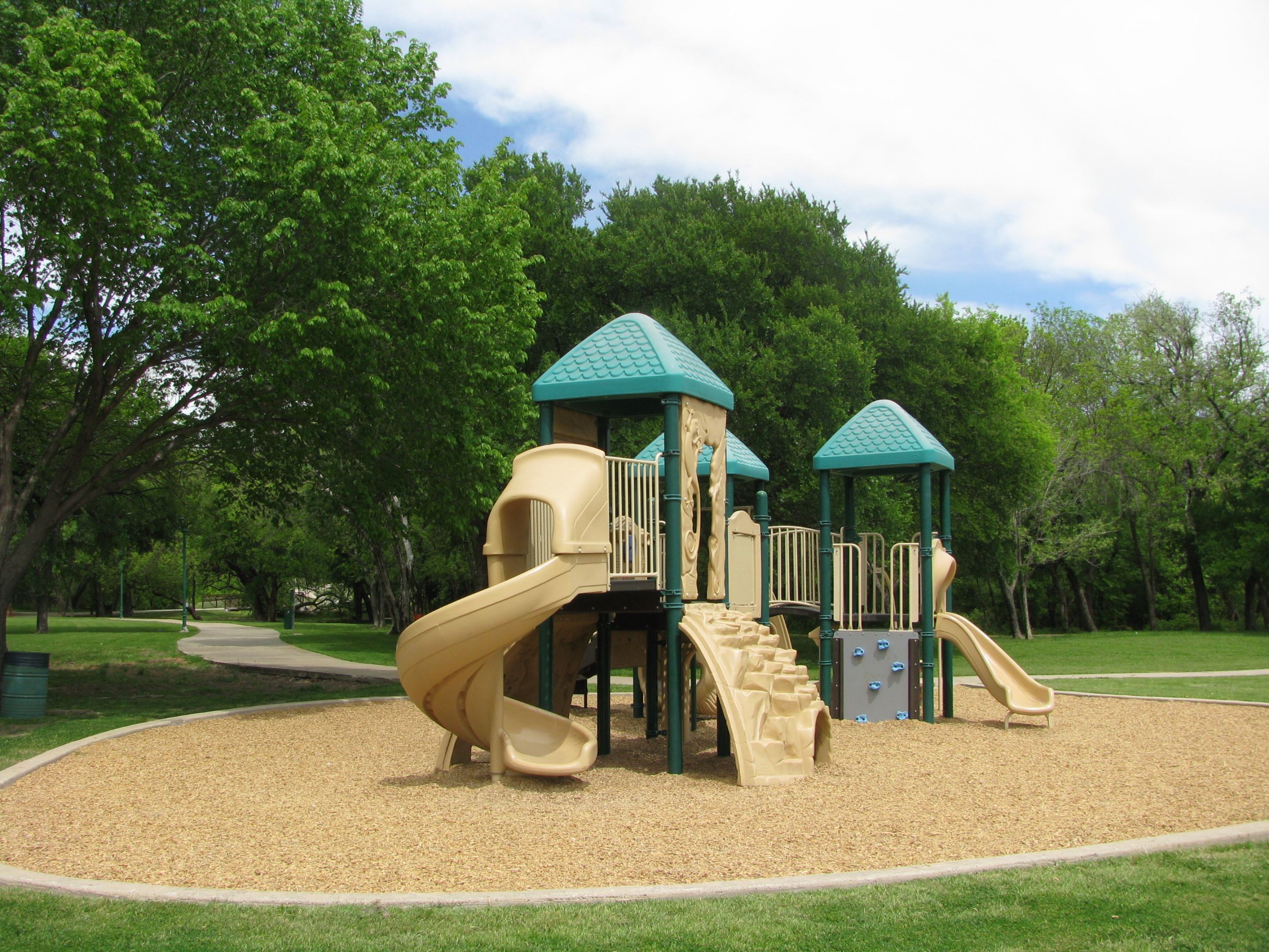 Playground with slides, climbing features and green-roofed towers, surrounded by trees
