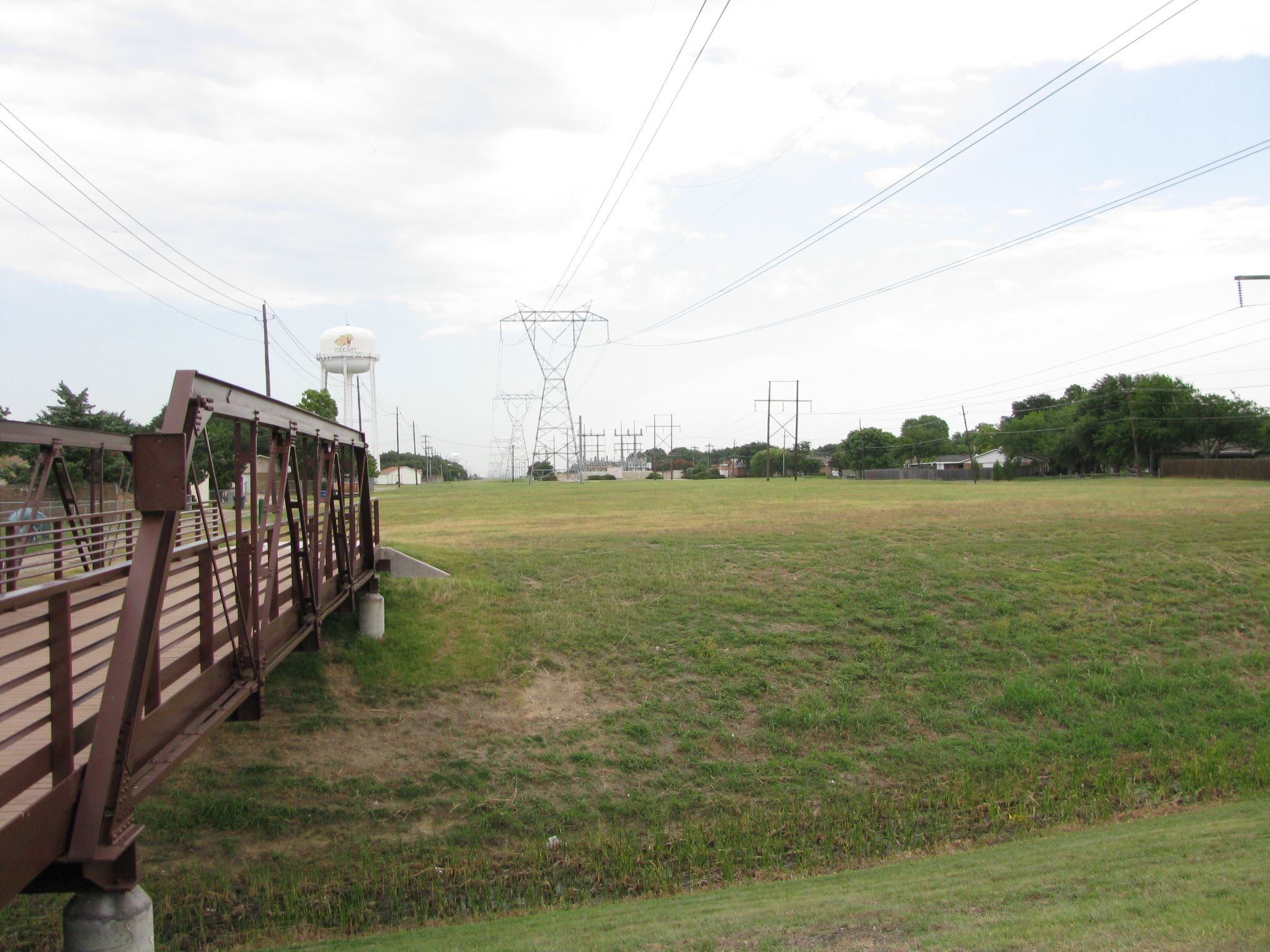 Pedestrian bridge over grassy area with power lines, water tower and houses in the distance