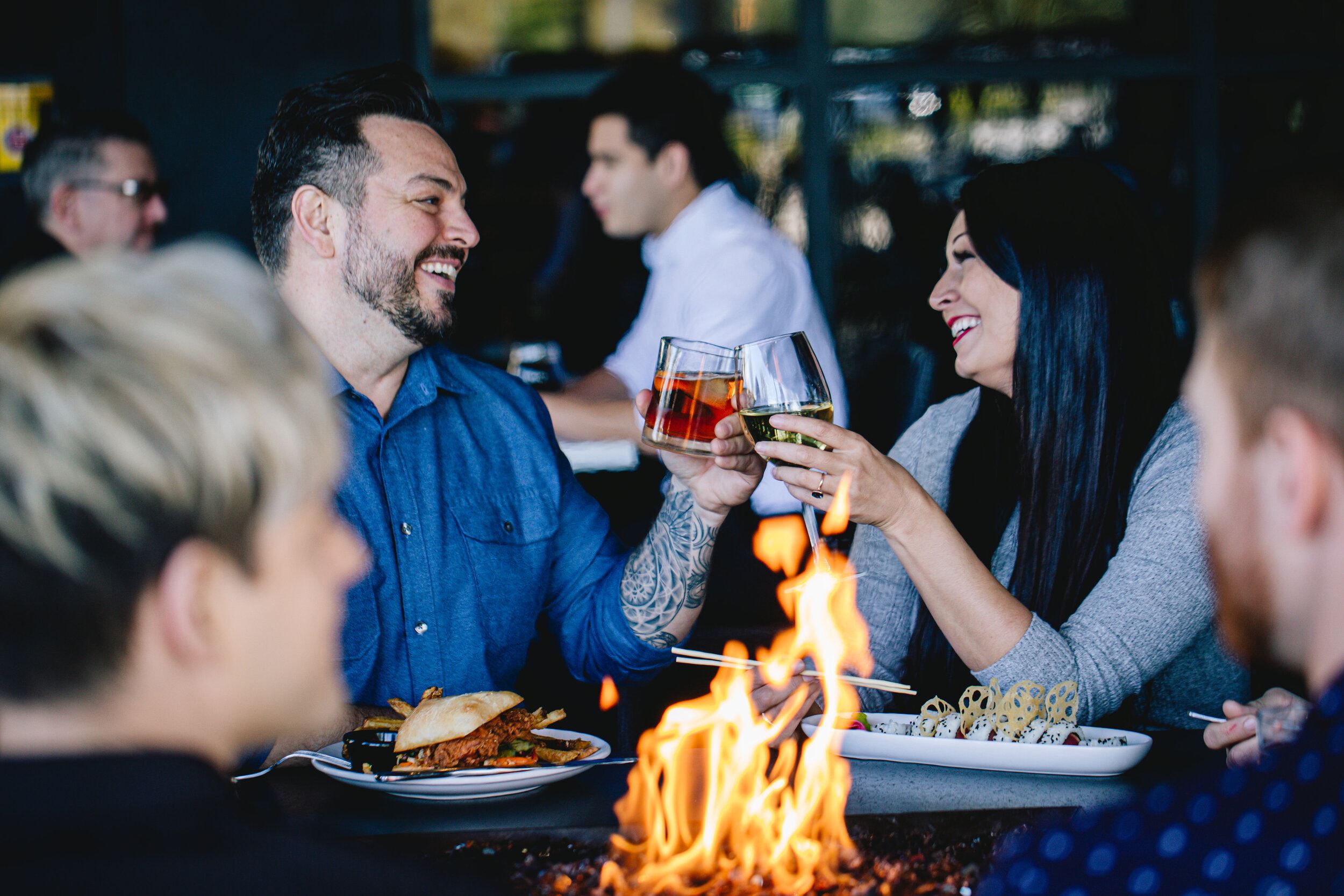 Smiling couple toasting drinks at a restaurant table with fire pit and food.