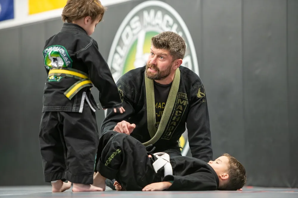 Jiu-jitsu instructor kneeling while teaching two young boys in black gis on a training mat.