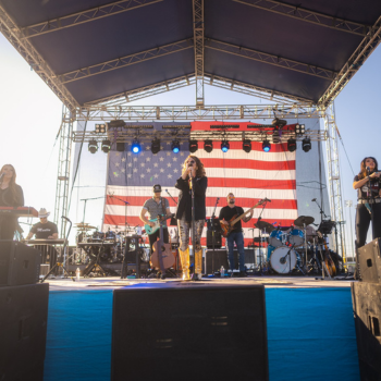 Band performing on outdoor stage with large American flag backdrop.