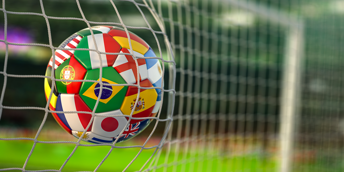 Soccer ball with international flags hits the goal net on a green field.